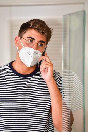 Focused looking young man wearing a face mask and glasses talking on his phone on an out of focus background. Communication and safety concept.の写真素材