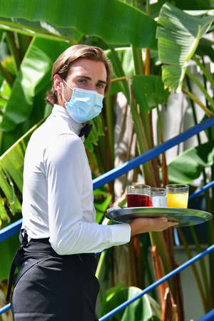 Young male waiter with a surgical mask holding a bar tray with assorted drinks outdoors on an out of focus background. Serving and safety concept.の写真素材