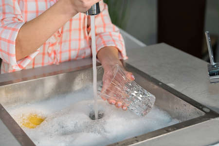 Close-up of a woman holding a glass and filling a kitchen sink with water from a faucet. Cleaning and hygiene concept.の写真素材