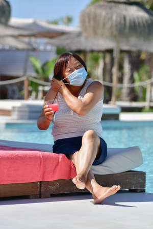 Asian woman holding a drink and fixing her surgical mask sitting at an hotel terrace on an out of focus background. New holidays and safety concept.の写真素材