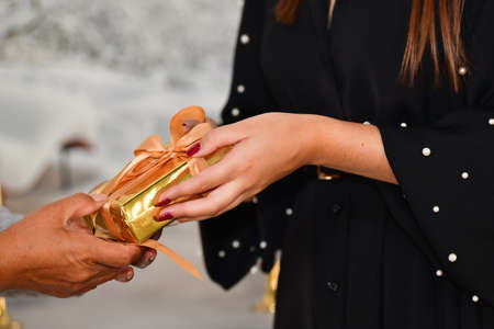 Close up of female hands giving a golden wrapped gift box to another female on an out of focus background. Christmas and gift giving concept.の写真素材