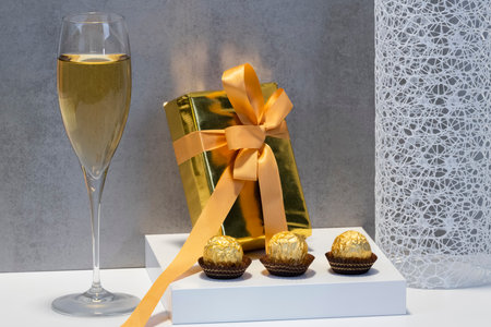 Selective focus of a golden wrapped gift box with wrapped chocolate sweets and a glass of champagne. Celebration and luxury concept.の写真素材