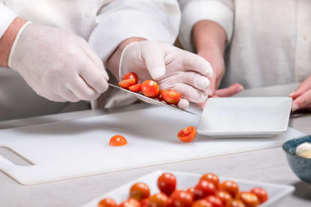 Male chef wearing gloves putting cut cherry tomatoes on a dish using his knife: selective focusの写真素材