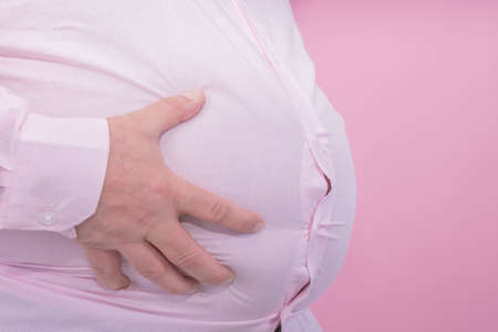 Fat man hand holding his big belly on a pink background: typical photo for beer belly: Selective focusの写真素材