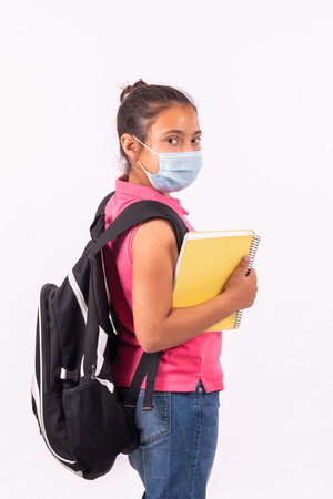 Little latina girl with a face mask carrying a big backpack looking back at the camera: School...