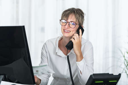 Front view of a businesswoman with eyeglass using computer and talking on the phone looking at cameraの写真素材