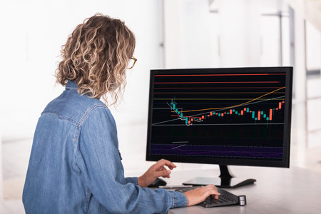 Rear side view of an unrecognizable woman in office using computer with statistics and charts on...