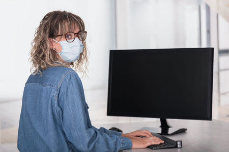 Rear view of an adult woman wearing protective face mask in office using computer with dark screen...