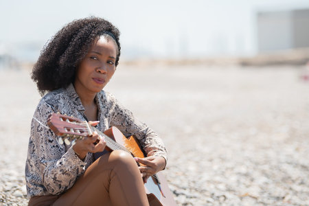 Young woman sitting on a gravel stone ground outside playing her acoustic guitar looking awayの写真素材