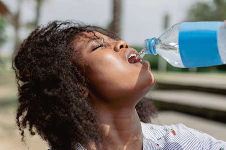Afro woman drinking water from a bottle while walking outdoors on a hot and sunny day.の写真素材