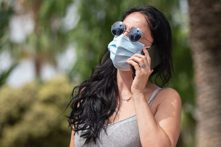 Woman in the park wearing surgical face mask and sunglasses talking with her phoneの写真素材