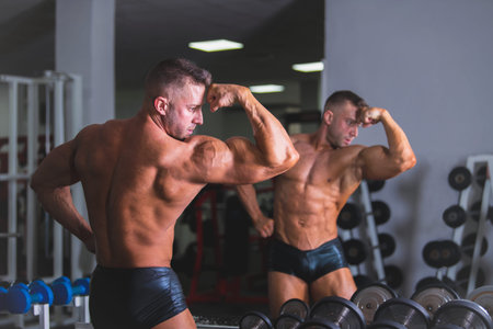 Bodybuilder man posing in front of a mirror after doing exercise at the gym.の写真素材