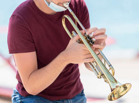 A young man with a surgical mask pulled down to play a trumpet. Music and new lifestyle concept.の写真素材