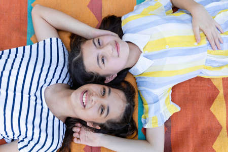 Top view of two girl children holding hands behind head and smiling while lying on the Bright and colorful floor clothes together. enjoy and looking at camera. Friends and friendship conceptの写真素材