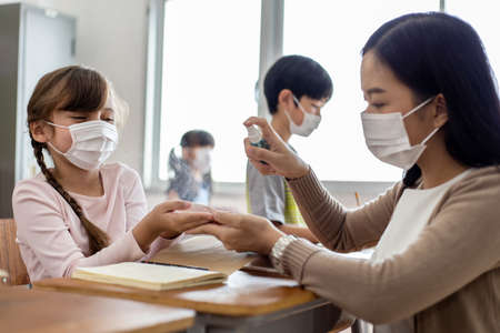 An Asian woman teacher wearing a medical flu mask. She injects alcohol spray onto the student's palm for Protect from Coronavirus (COVID-19) Outbreak - Healthcare Concept.の写真素材