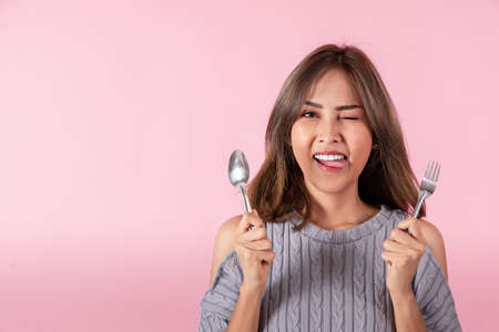 Portrait of an Asian woman holding a spoon and fork in his hand. She is choosing a food menu. Shot of the studio. 
Separately on the pink background.の写真素材