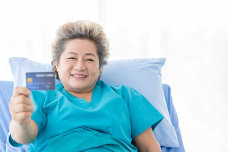 An asian old woman in bed in a hospital Smiling and showing credit cards with a white background...