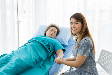 Asian families visit critically ill elderly people at the hospital. A woman holds the hand of a sleeping mother in a hospital bed. The concept of love of family and health insuranceの写真素材