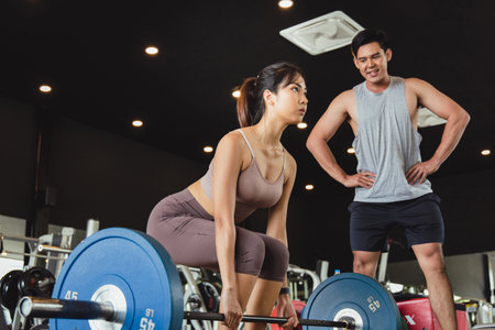 Asian women in sportswear preparing to lift barbells in fitness gym. Close up of young woman doing deadlift  workout with her handsome muscular personal trainer. Concept Exercise  for good shapeの写真素材