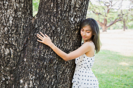 A beautiful Asian teenage girl hugs a large tree in the forest with love. Her face is cheerful....