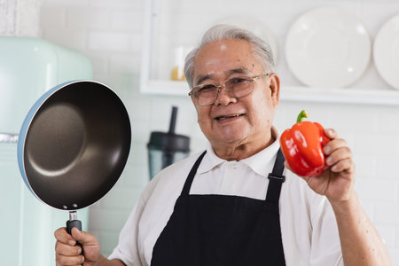 Portrait of an elderly Asian man standing in the kitchen He holds the pan and vegetables in his hand. Grandfather looked at the camera and smiled. He prepares healthy foodの写真素材