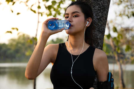 athletic young women after run. women resting after jogging at the park. Young women wear headphones to listen to music while jogging. female drinking water from bottle after workoutの写真素材
