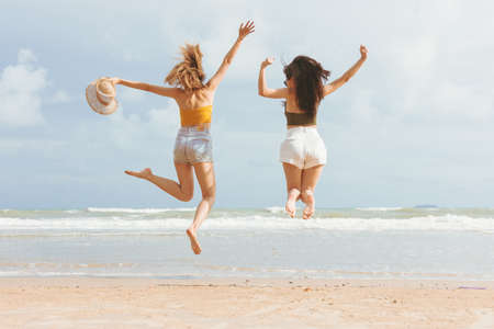 Two Asian women in summer casual clothes Jumping on the Beach Having Fun. Young Female friends wearing sunglasses, smiling having fun on vacation in tropical country. back view.の写真素材