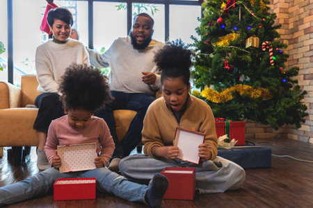 Two young girls are thrilled and surprise at the Christmas presents their father and mother give. Daughter sitting opening gift box. African American family unpacking Christmas giftの写真素材