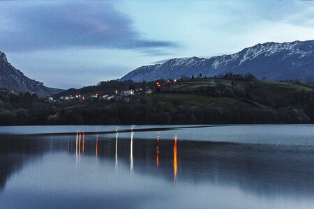 Landscape of a lake at dusk with a small town on the other shore and the reflection of lights in the water. Snowy mountains in the background in Asturias.の写真素材