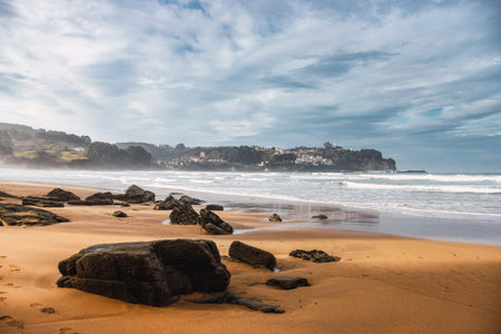 Rocks in the sand on the beach in the foreground during a sunny and cloudy day in northern Spain. Relating to tourism, travel, oceans and nature in Asturiasの写真素材