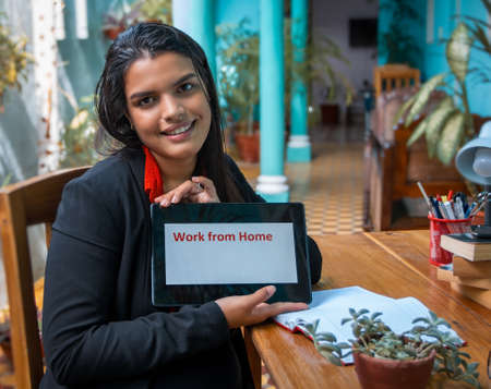 An smiling young woman sitting at a table with a tablet in her hands. On the tablet screen is written Work from home in red lettersの写真素材