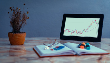 Screen of a tablet on a desk with a chart of currency pairs. On the desk there is an out of focus...