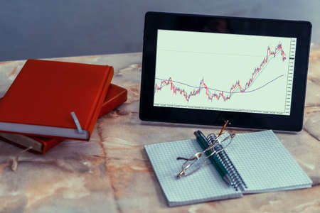 A tablet screen on a desk displaying a currency pairs chart. There are a notebooks, a pen and a glasses too.の写真素材