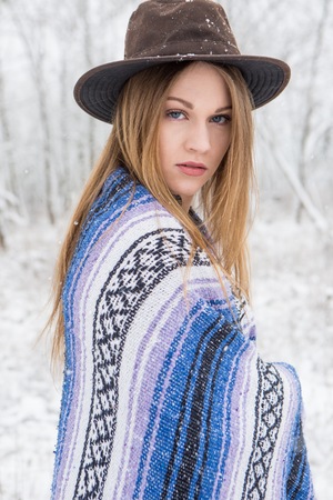Young adult woman with hat and wrapped in a blanket standing outdoors in the snow.の写真素材