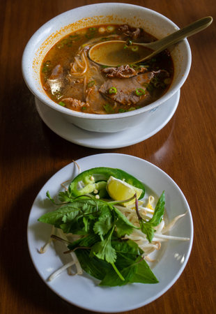 A hot bowl of Spicy Beef Pho with vegetables and herbs on the side. Vietnamese street food that has...