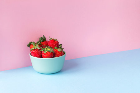 Minimalist image of a bowl full of strawberries on a light blue and pink background. Copy space...