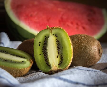 Close-up of a group of kiwis, one of them cut in half. Blurred background with a red watermelon. On a white tablecloth with blue lines.の写真素材