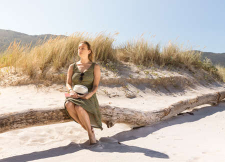 A middle-aged blonde woman sitting thoughtfully on a tree trunk at the beach with a hat and a book on her legsの写真素材