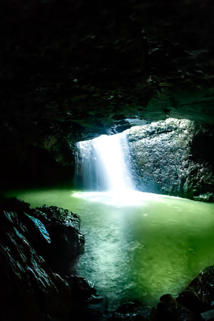 Shiny waterfall flows in the dark stone cave that brighten with the sunlight, the water turns green because of the lightの写真素材