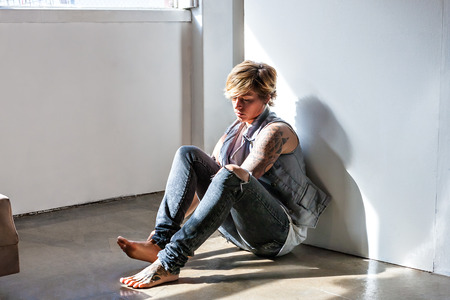 Sad looking young woman with short haired sitting on the floor with a tattoo on her arm and lean on the white wallの写真素材