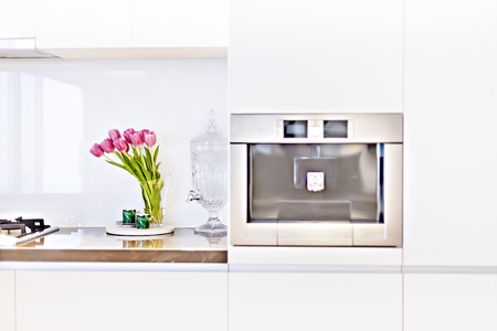 Pink rose flowers on the shiny counter top in the kitchen with a glass jug water filter and store beside the blurred oven, there are green leaves of the plant beside small mugsの写真素材