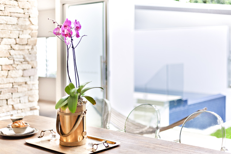 Purple orchid flowers in a gold color vase close up on the wooden table, there are a brass tray and some foods on mugs, background is blurred and white which illuminated with sunlightの写真素材