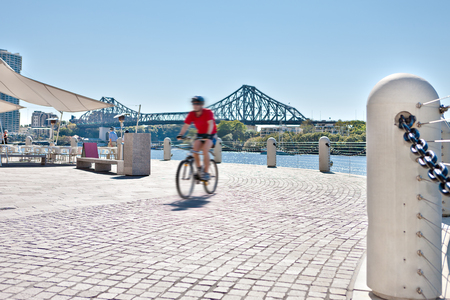 man riding a bicycle on the riverside stone tiles street with a concrete pillar fence that attached with steel chain fixed to the stone tile road and Brisbane City and buildings from the background from Queensland, Australia.のeditorial素材