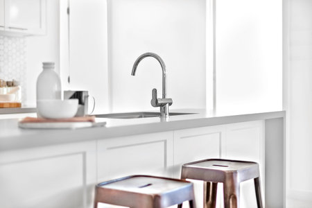 Modern kitchen counter focusing the silver tap and blurred beside a black and plastic chairs near an...