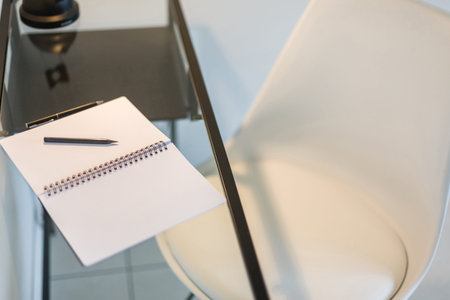 Top view of a stylish white chair and a glass study table with a notebook, pen and pencil kept on it