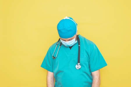 doctor dressed as a surgeon in green with stethoscope and mask on a yellow background with a worried expression.の写真素材