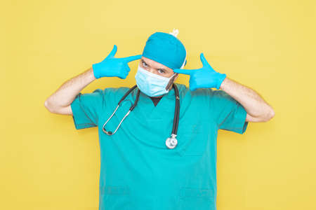 doctor dressed as a surgeon in green with stethoscope and mask on a yellow background with a worried expression.の写真素材