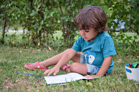 Young Caucasian school boy sitting in park outdoor doing school homework. Child kid writing in notebook with pencil outside. Self education learning studying. Early development for children.の写真素材