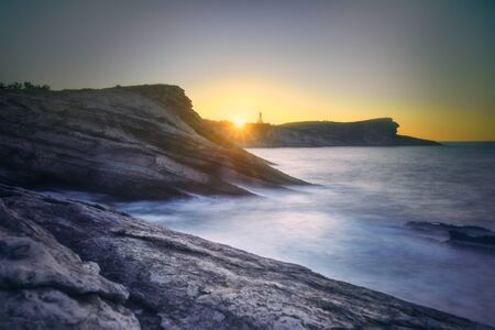 Stormy sea waving near rough rocky shore against bright sundown sky in evening in natureの写真素材