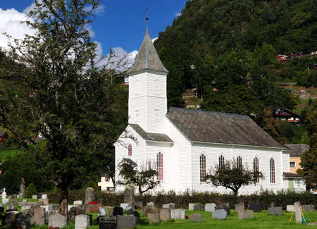 White Wooden Church With A Cemetery In The Foreground In Oystese Surrounded By Large Mountains On A Sunny Summer Day With A Clear Blue Sky And A Few Cloudsの写真素材